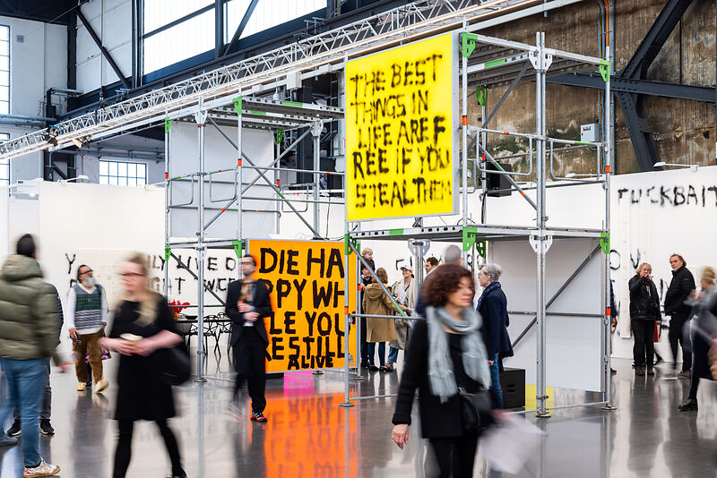Messehalle mit Gerüst-Installation, an der großformatige Text-Tafeln in Gelb und Orange hängen.
